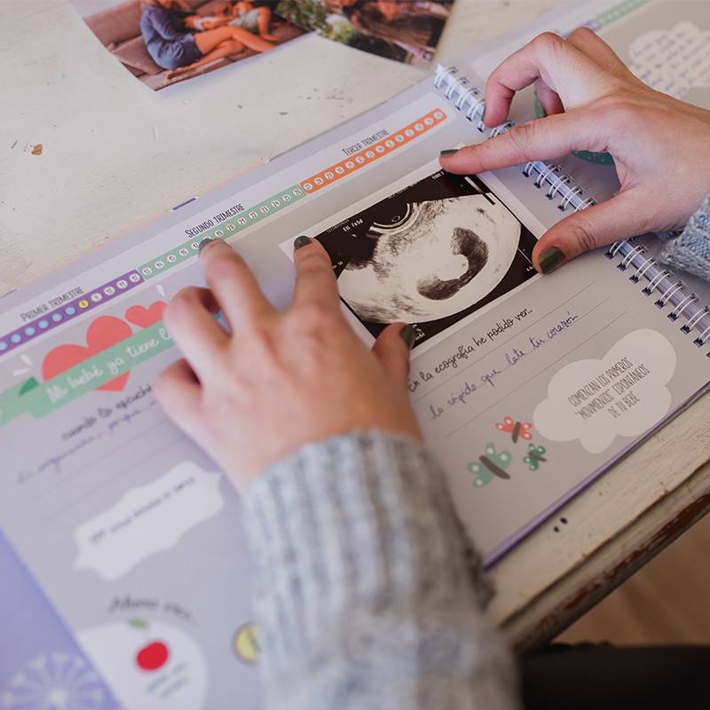 Primer plano de manos colocando una foto de ultrasonido en un diario de embarazo decorado con ilustraciones.